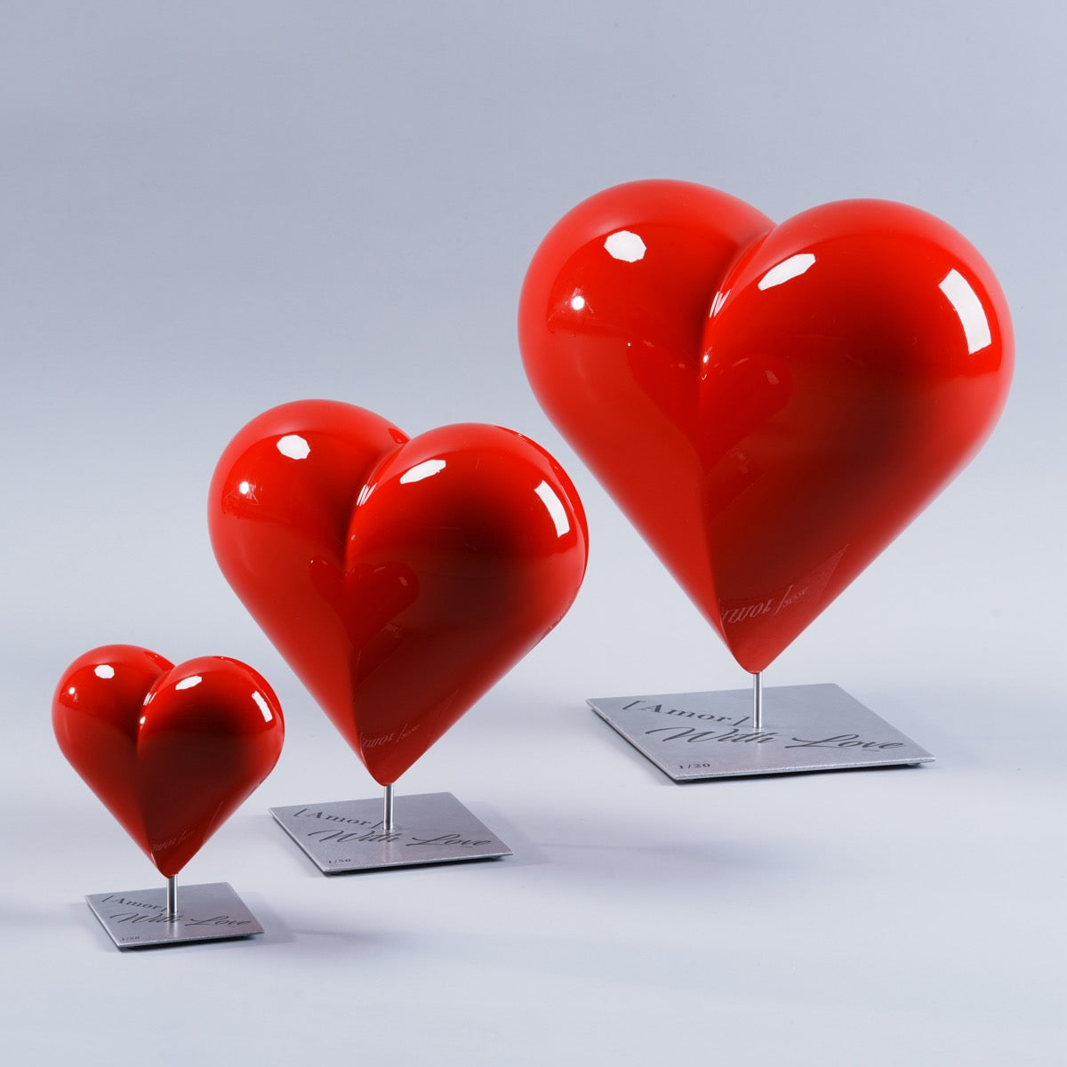 Trois sculptures d’art HE[ART]™ en forme de cœur rouge laqué brillant, disposées en diagonale et présentées en trois tailles différentes. Chaque cœur est monté sur une tige métallique et repose sur un socle carré en acier inoxydable gravé « [Amor] With Love 2026 », photographié en studio sur fond gris clair.