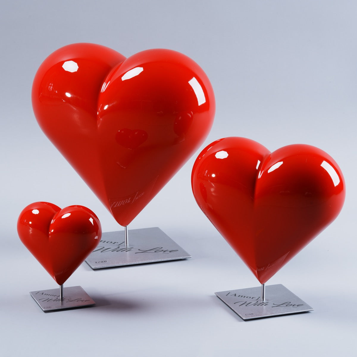 Ensemble de trois sculptures d’art HE[ART]™ en forme de cœur rouge laqué brillant, présentées en trois tailles différentes. Chaque cœur est fixé sur une tige métallique centrale et repose sur un socle carré en acier inoxydable gravé « [Amor] With Love 2026 », photographié en studio sur fond gris clair.