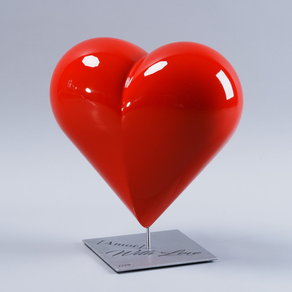 Sculpture d’art en forme de cœur rouge laqué brillant, modèle HE[ART]™, édition limitée Saint-Valentin par l’artiste Amor. Cœur posé sur une tige métallique fixée à un socle carré en acier inoxydable gravé, photographié sur fond gris clair en studio.