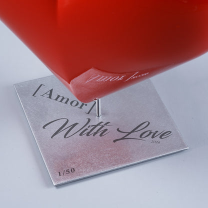 Gros plan de la base d’une sculpture en forme de cœur rouge laqué HE[ART]™, montrant la pointe du cœur fixée sur une tige métallique. Socle carré en acier inoxydable avec gravure laser « [Amor] With Love 2026 » et numérotation 1/50, photographié sur fond gris clair en studio.